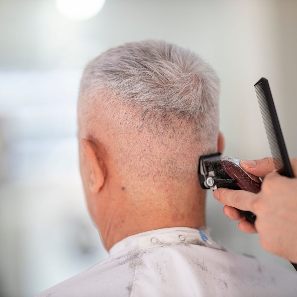 Elderly man getting hair trimmed by barber indoors using clipper and comb.